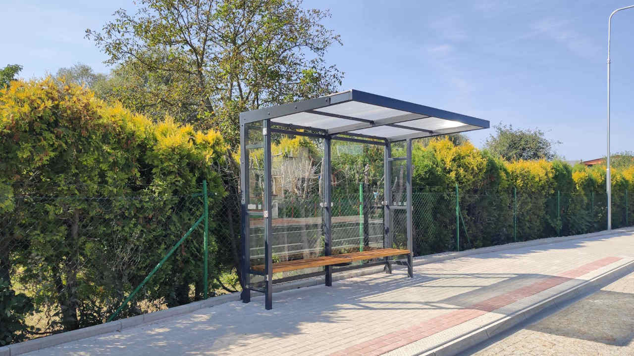 Autobusová zastávka typ TECHNIK s plochou střechou zadní stěnou ze skla  a lavičkou ze dřeva v  prostředí se živým plotem